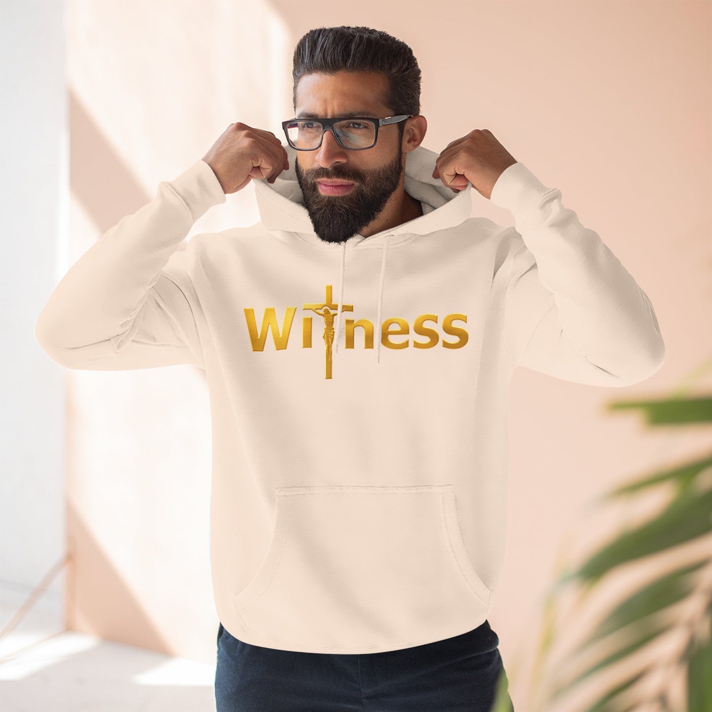 Light-colored pullover hoodie with gold “Witness” print and cross emblem, hood and pocket.