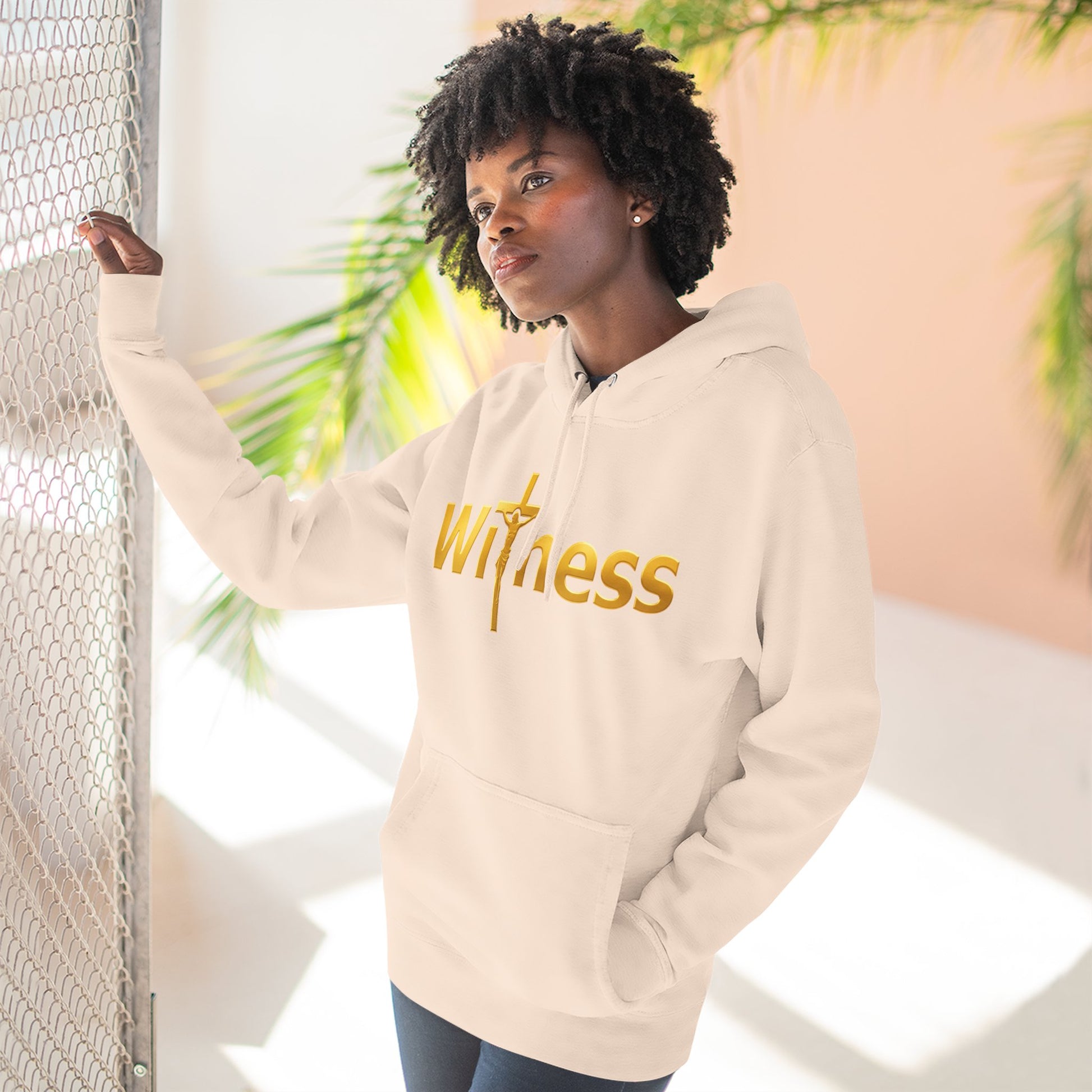 Light-colored pullover hoodie with gold “Witness” print and cross emblem, hood and pocket.